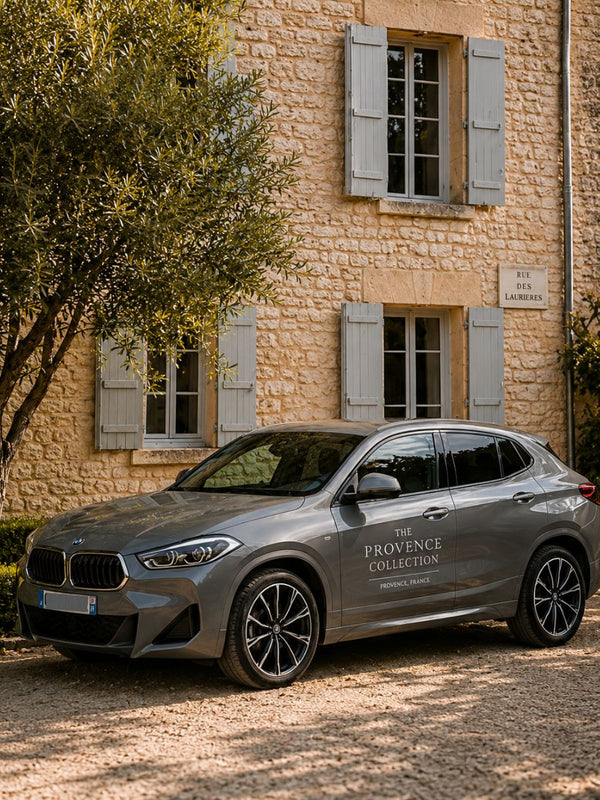 Gray BMW SUV with The Provence Collection logo parked by a stone house with pale blue shutters and leafy trees.