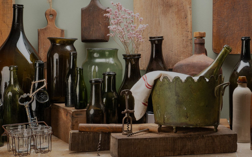 Vintage green and brown glass bottles with corkscrews, glasses, metal pot, and dried flowers on rustic wood table.
