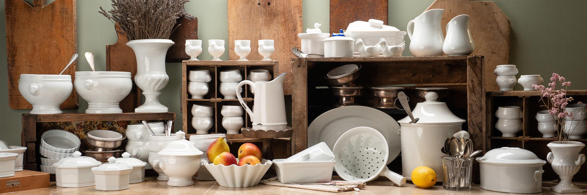 Vintage white ceramic dishes, bowls, pitchers, and serving pieces displayed on wooden crates with fruit and dried flowers.