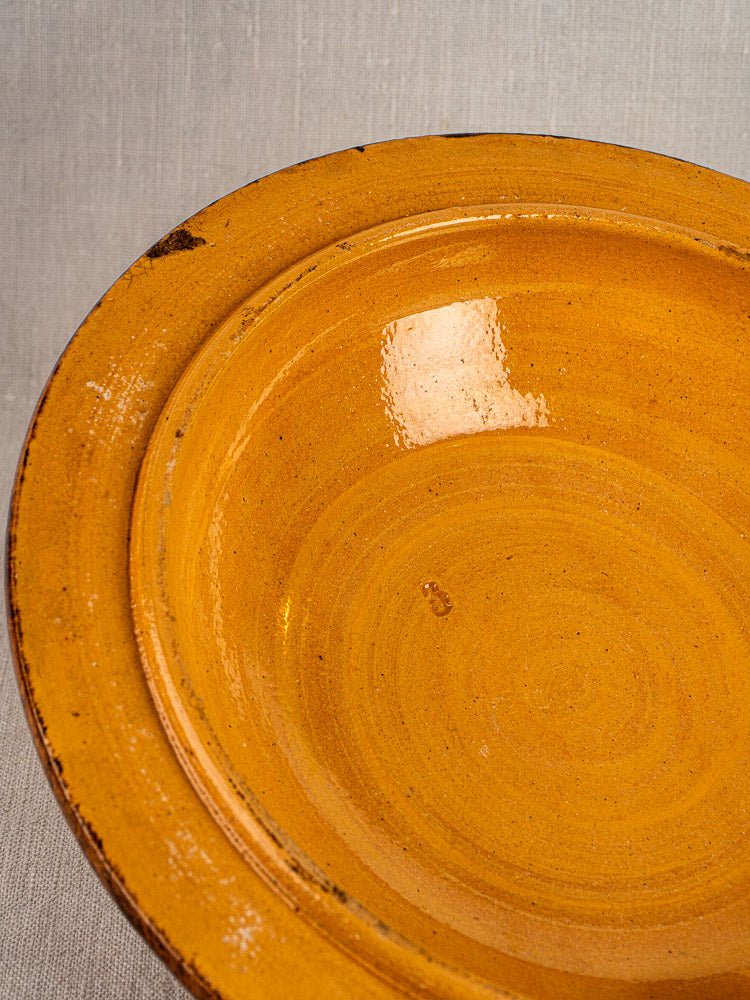 Round yellow-orange glazed ceramic bowl with dark rim spots, Alsace terracotta, on neutral fabric.