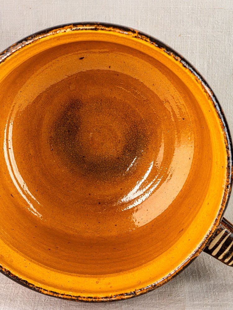 Round orange glazed earthenware bowl with handle from Alsace, on light gray textured surface.