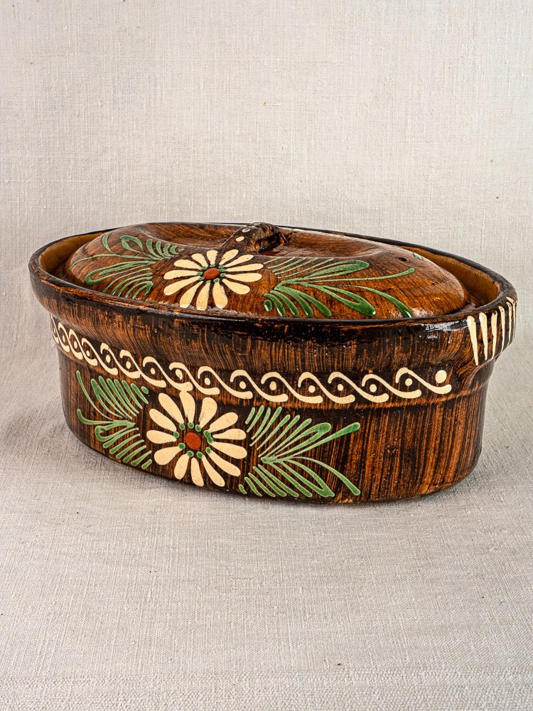 Alsace-style glazed terracotta oval dish with lid, floral slip decoration on neutral fabric background.