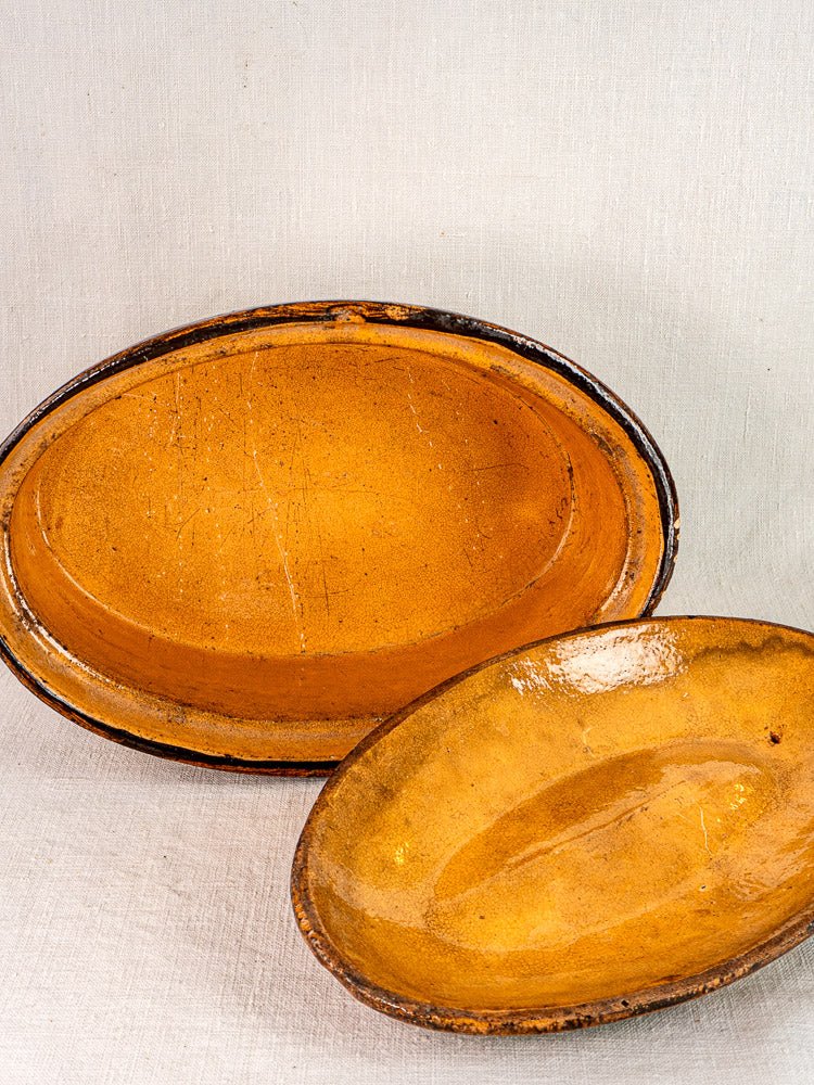 Pair of oval Alsace glazed terracotta dishes with yellow-brown slip decoration on light textured surface.