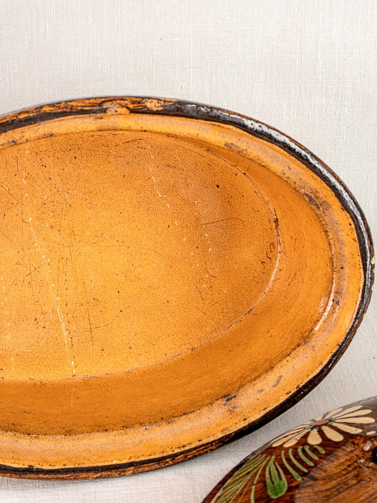 Large oval Alsace glazed terracotta cookware with worn yellow-orange interior and dark rim; smaller floral bowl partially visible.