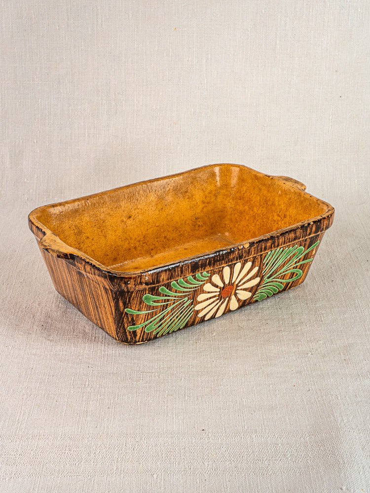Rectangular French terracotta baking dish with brown glazed interior, floral slip decoration, on neutral fabric.