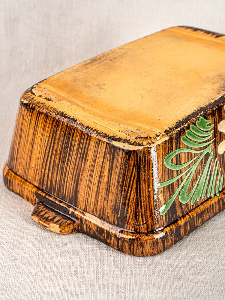 Alsace glazed terracotta cookware with woodgrain motif, brown edges, yellow top, green accents on beige fabric.