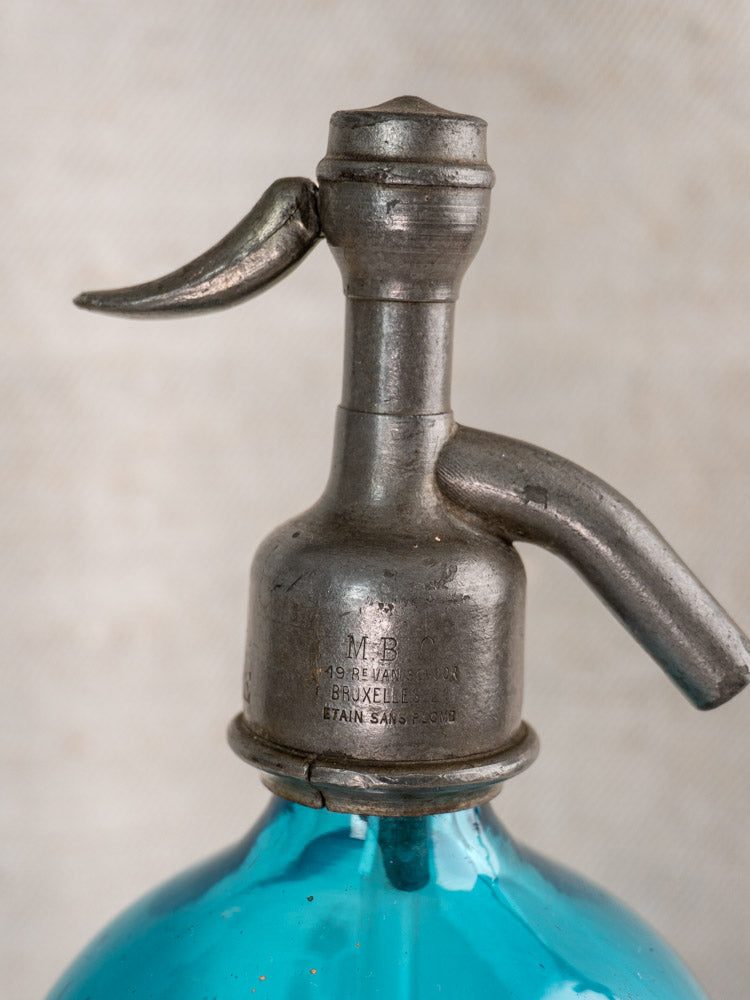 Antique Belgian soda siphon with metal top and turquoise blue glass, Bruxelles etched, on a neutral textured background.