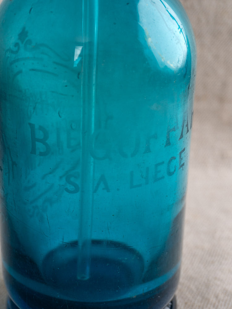 Antique Belgian soda siphon in turquoise blue glass with faint Bruxelles etching, set on a neutral textured surface.
