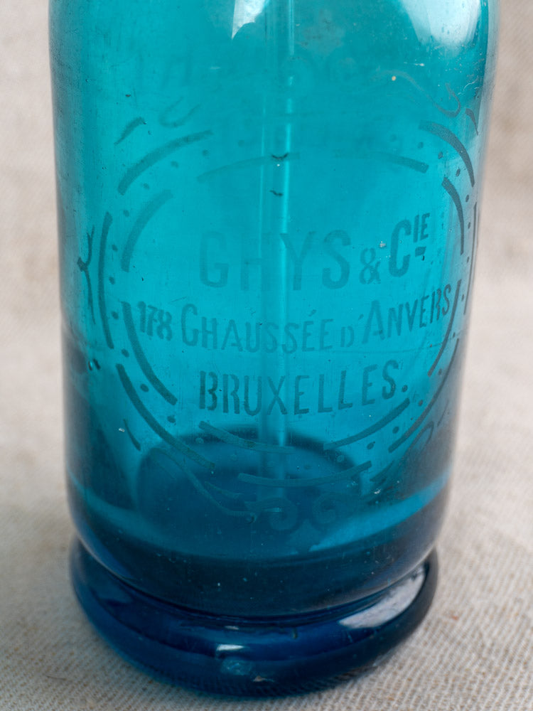 Antique turquoise blue Belgian soda siphon with "Bruxelles" etching, set on a neutral fabric background.