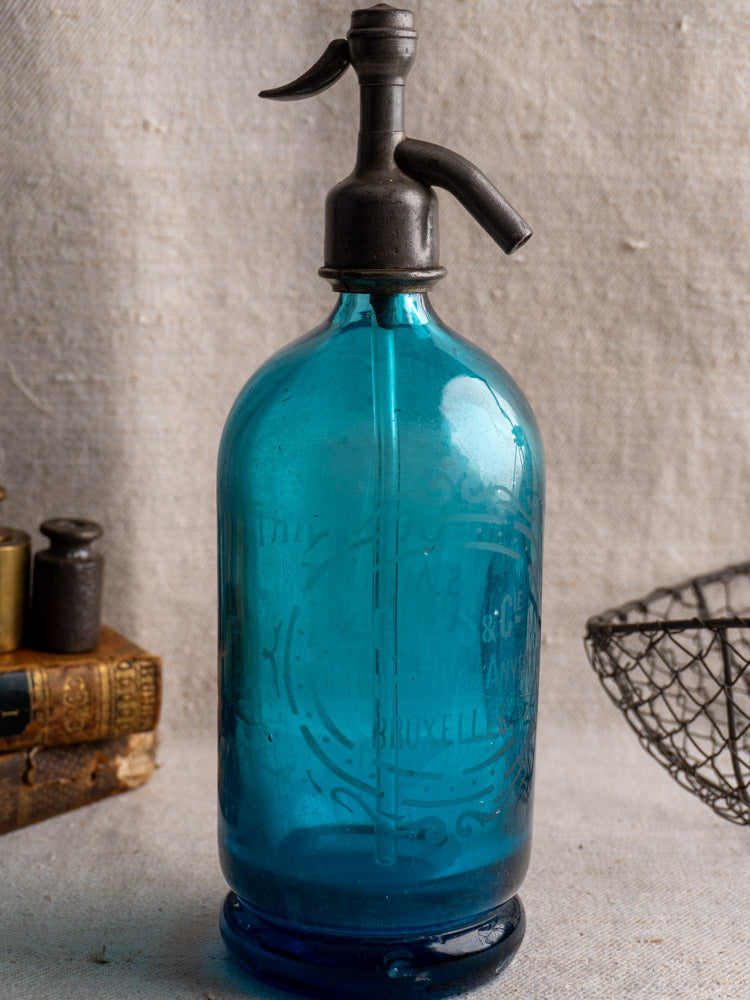 Antique Belgian soda siphon in turquoise blue glass with "Bruxelles" etching and metal top on textured beige fabric.