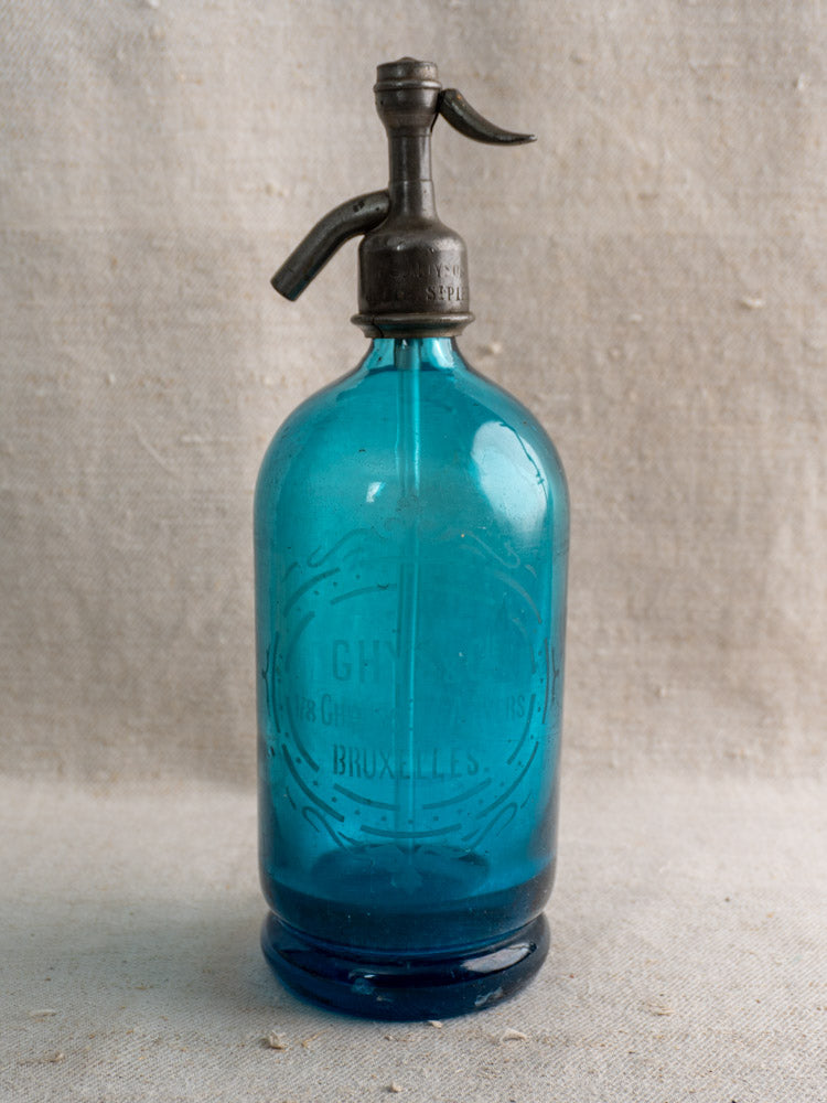 Antique Belgian soda siphon in turquoise blue glass with "Bruxelles" etching and metal top on textured beige fabric.