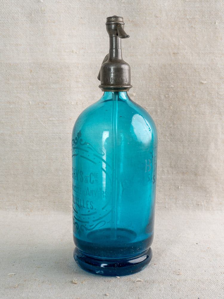 Antique Belgian turquoise blue glass soda siphon with “Bruxelles” etching, set before textured beige fabric.