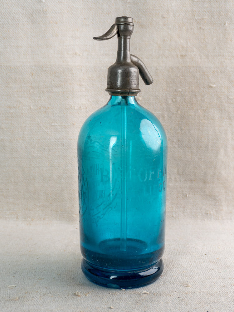 Antique Belgian soda siphon in turquoise blue glass with Bruxelles etching on a neutral textured fabric background.