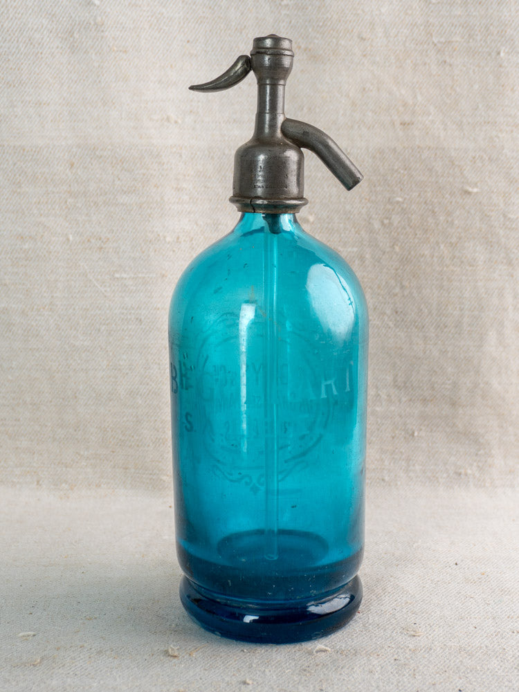 Antique Belgian soda siphon in turquoise blue glass with "Bruxelles" etching and metal top on neutral fabric.