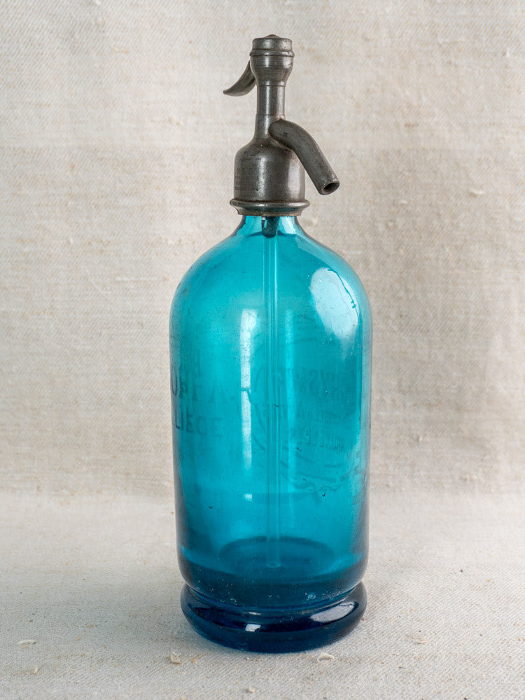 Antique Belgian soda siphon in turquoise blue glass with 'Bruxelles' etching, metal top, on neutral surface with light fabric background.