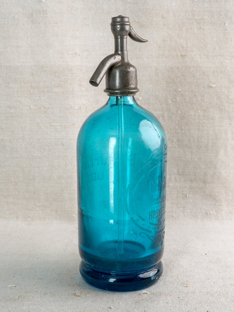 Antique Belgian soda siphon in turquoise blue glass with "Bruxelles" etching and metal spout on light textured fabric.