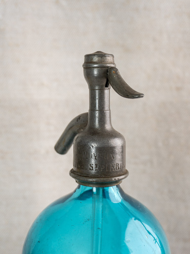 Antique Belgian soda siphon in turquoise blue glass with "Bruxelles" etching, metal top and spout, neutral textured backdrop.