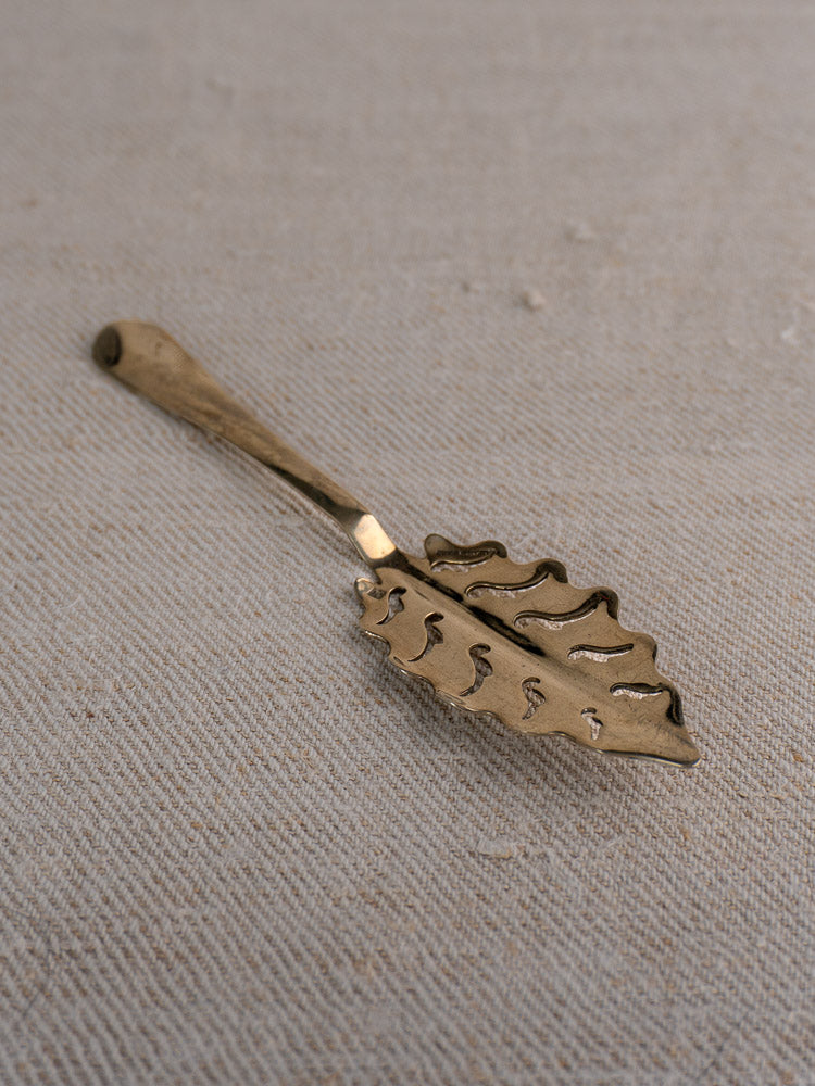 Gold-toned Cailar Bayard absinthe spoon with leaf pattern detailing, displayed on textured beige fabric.