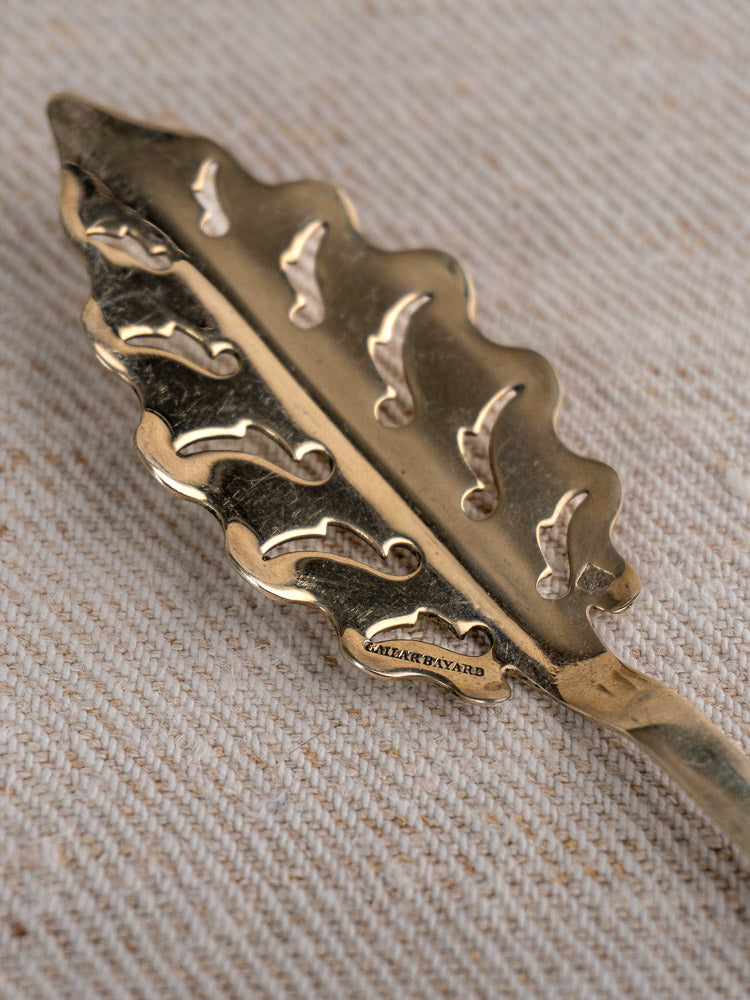 Gold leaf-patterned antique French absinthe spoon from Cailar Bayard on textured beige fabric with ATELIER DEVARD label.