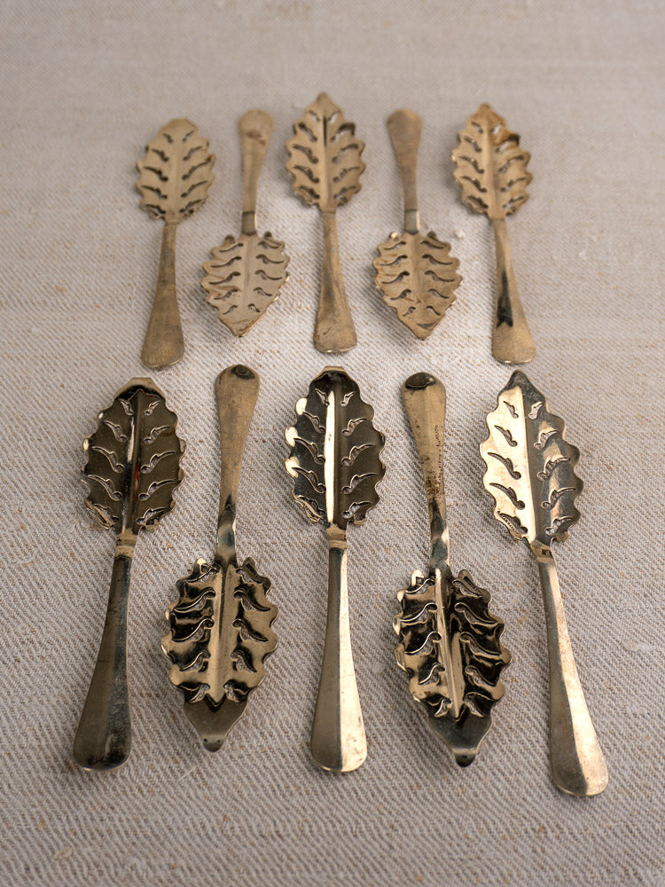 Set of 10 antique French absinthe spoons by Cailar Bayard with leaf pattern handles, displayed on beige fabric.