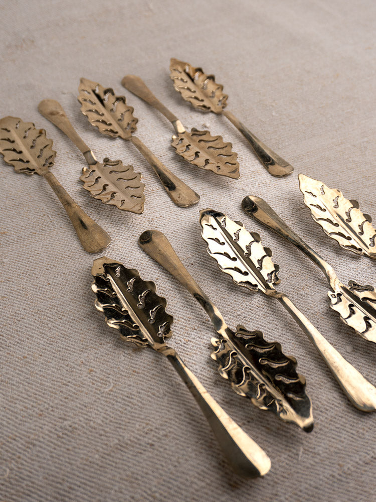 Set of 10 antique French absinthe spoons by Cailar Bayard with leaf pattern, displayed on beige textured fabric.