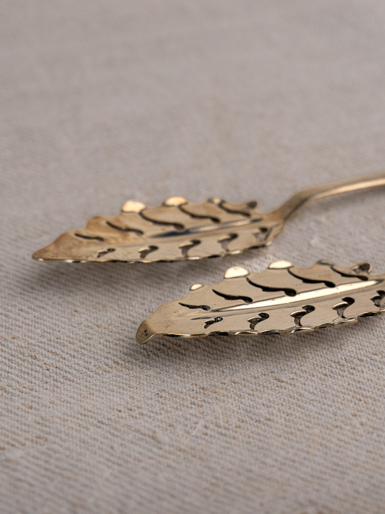 Set of ten antique French absinthe spoons by Cailar Bayard with leaf cut-out pattern on beige textured fabric.