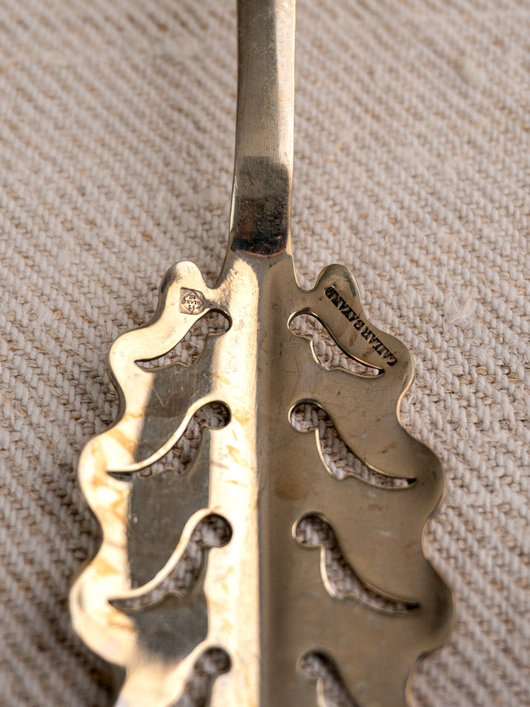 Gold-toned Cailar Bayard absinthe spoon with wavy edges, leaf pattern, on textured beige fabric.