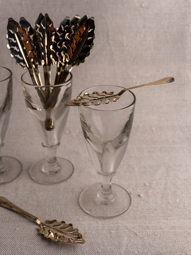 Set of 10 antique French absinthe spoons by Cailar Bayard with leaf pattern, displayed in glass goblets on a light textured surface.