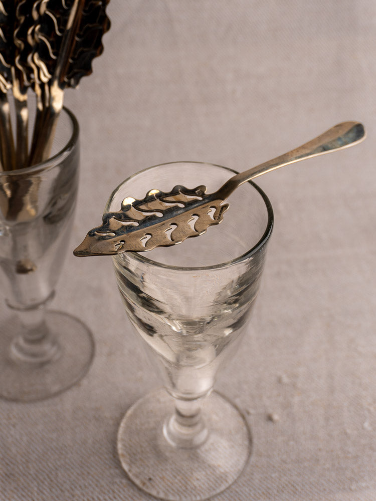Set of 10 antique French Cailar Bayard absinthe spoons with leaf motifs, displayed on a vintage glass goblet.
