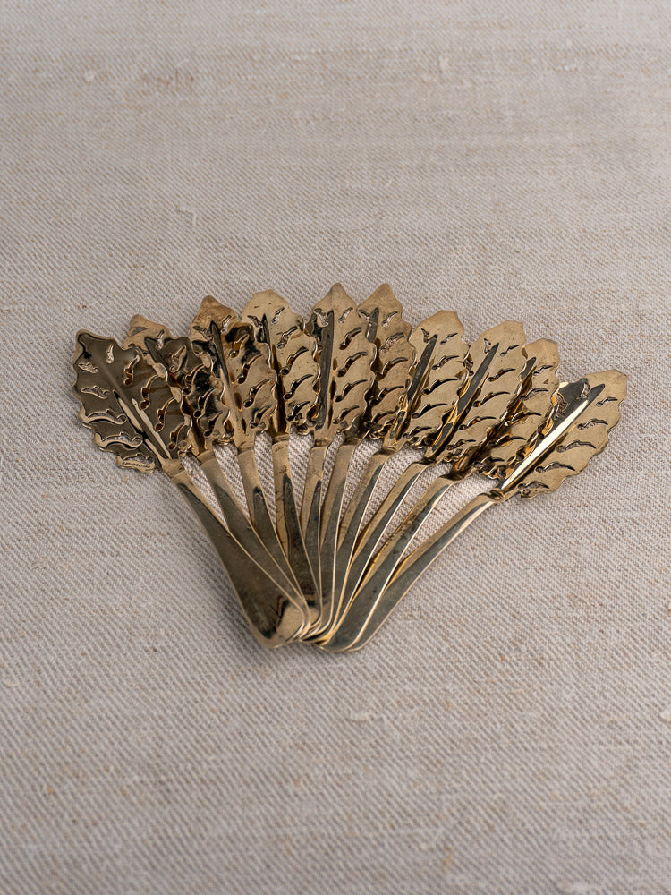 Set of 10 antique French Cailar Bayard absinthe spoons with leaf pattern, fanned on textured beige fabric.