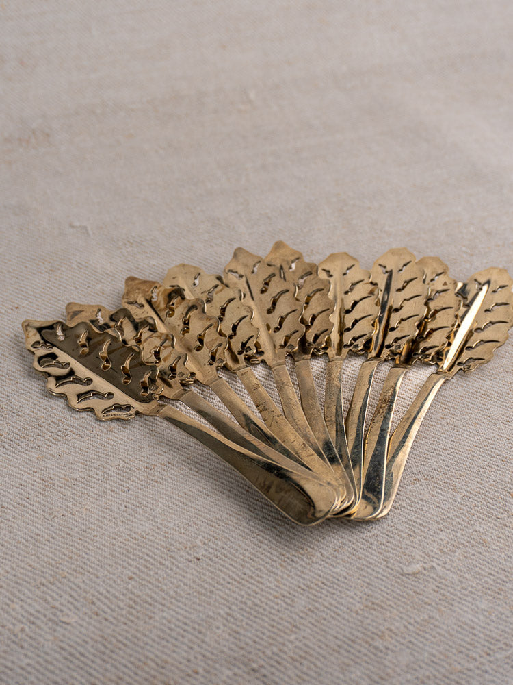Set of ten antique French absinthe spoons by Cailar Bayard with leaf-patterned handles on a neutral textured fabric.
