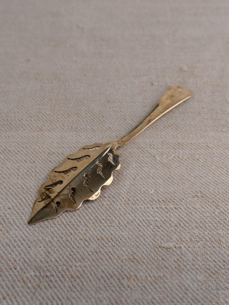 Set of 10 antique French Cailar Bayard absinthe spoons with leaf motifs on textured beige fabric.