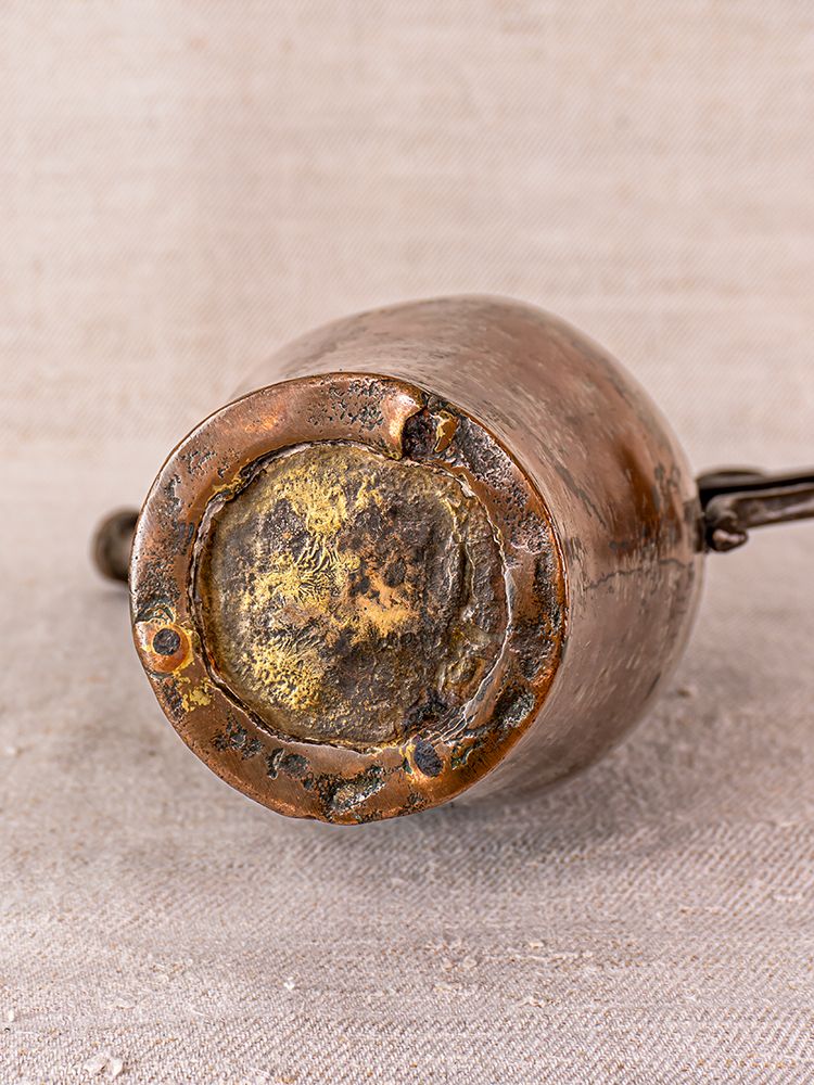 Worn, tarnished bottom of 19th-century Anatolian tinned copper ibrik with scratches and rivets on neutral fabric.
