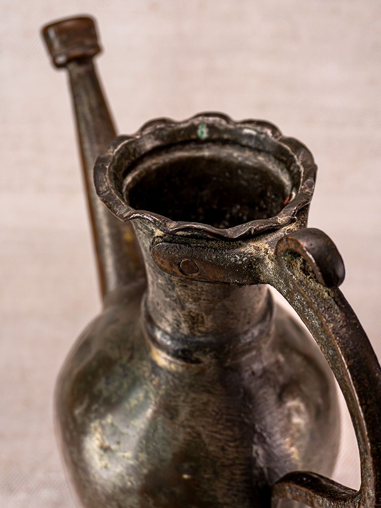 Antique Anatolian tinned copper ibrik watering can with scalloped rim and curved handle, neutral textured background.