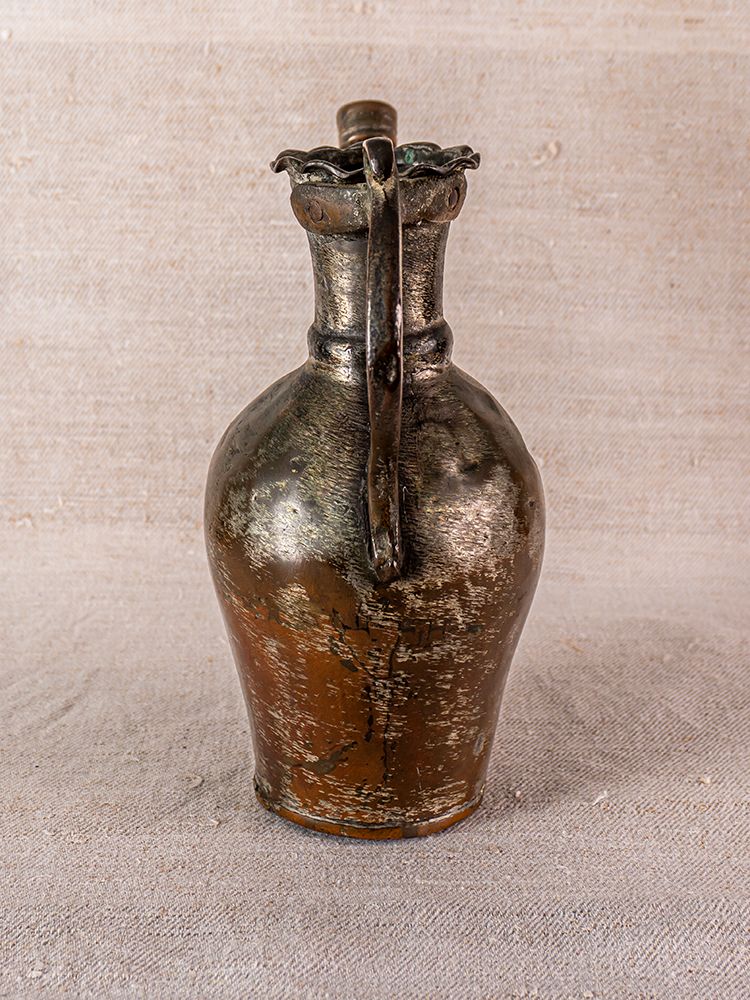 Antique Anatolian tinned copper ibrik watering can with weathered finish and neck handle on textured beige fabric.