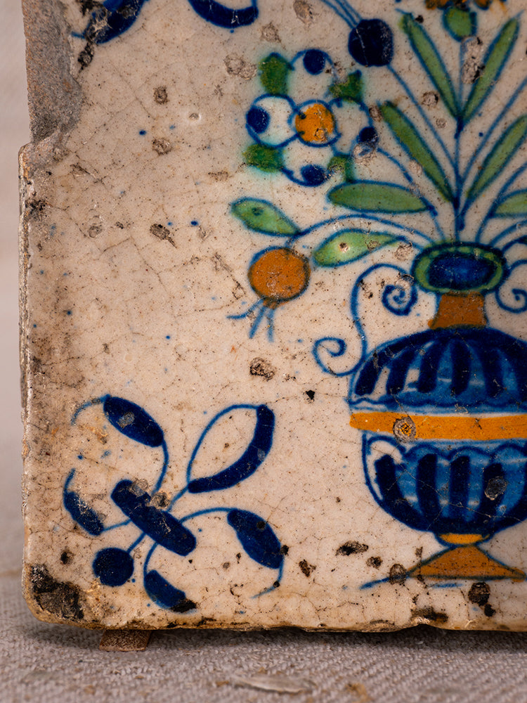 Antique French faience tile from Provence with blue vase and orange, green stylized flowers on cream weathered background.