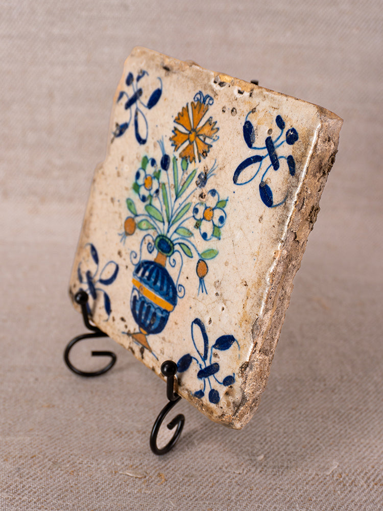 17th century French faience tile from Provence with floral vase motif on black metal stand, beige fabric backdrop.