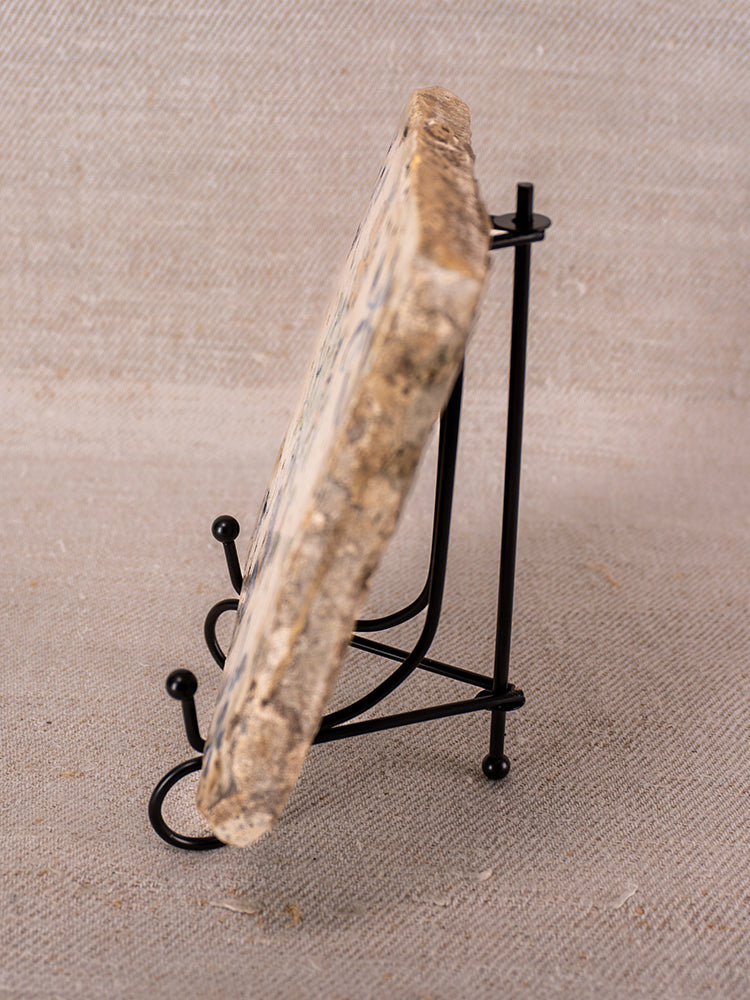 Antique French faience tile with polychrome flower vase, displayed on black stand, beige fabric background, visible rough edges.