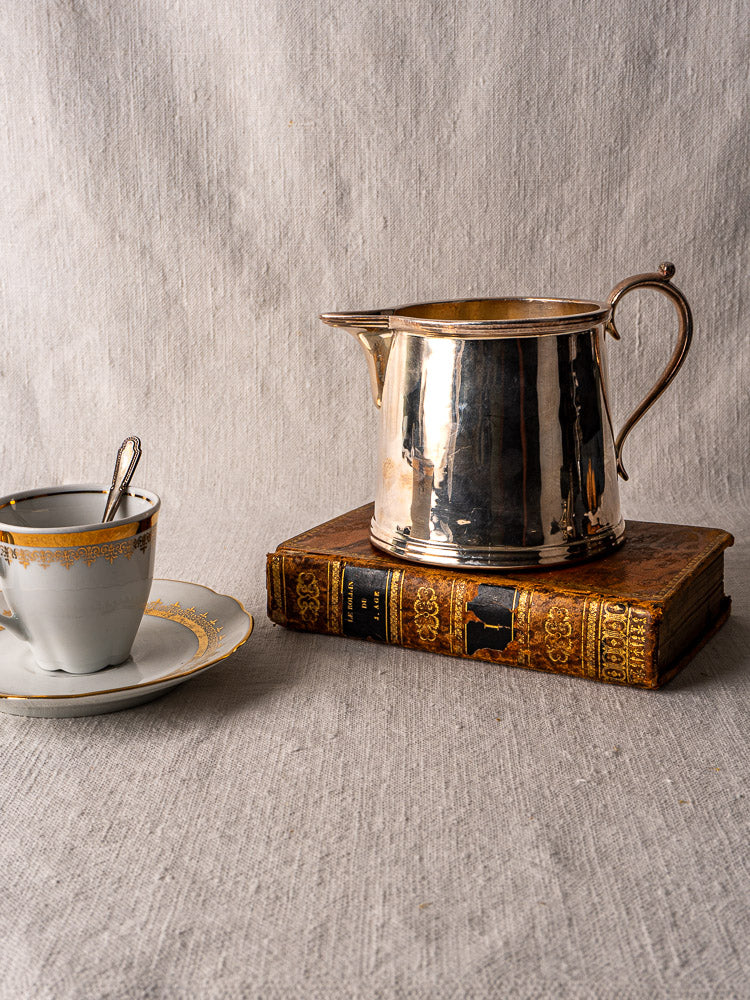 Antique English silver plate milk jug by Walker & Hall on vintage book with china cup and saucer, gray fabric background.