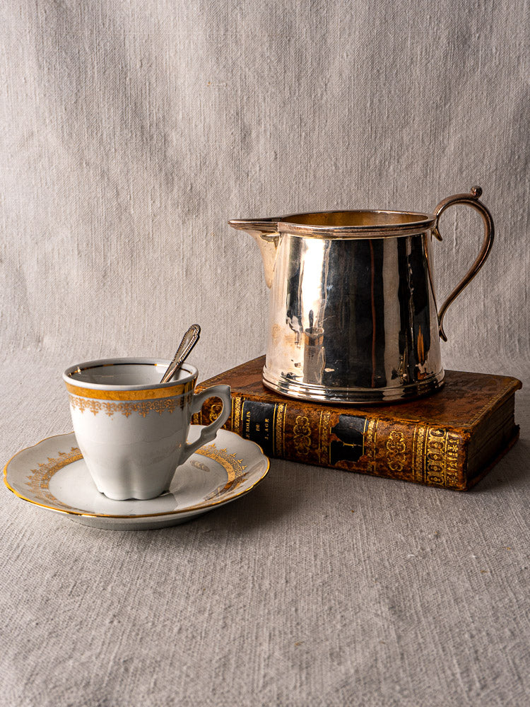 Vintage porcelain teacup and saucer with gold trim, silver plate milk jug, and ornate book on textured cloth.