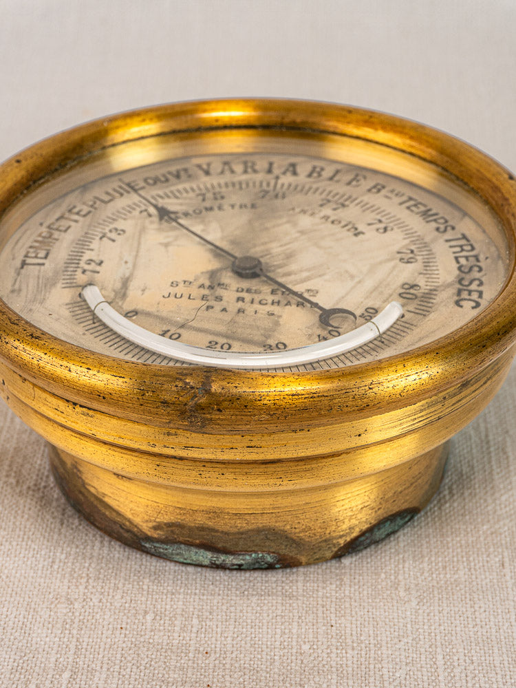 Antique French brass barometer by Jules Richard Paris with weathered patina on beige fabric surface.