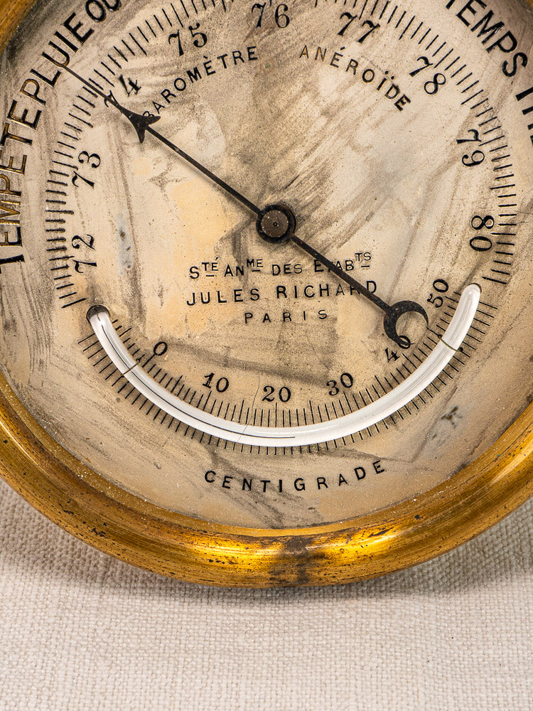 Antique French barometer by Jules Richard Paris in brass case with worn French dial, black pointers, on light fabric surface.