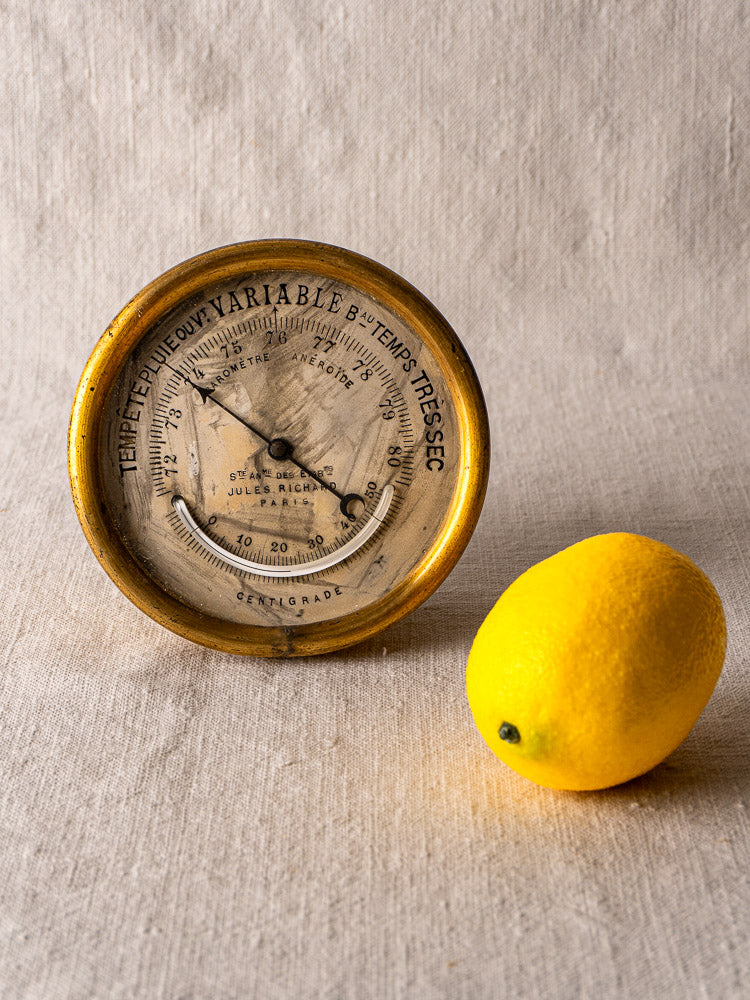 Antique French brass barometer by Jules Richard Paris with weathered dial on beige fabric beside a yellow lemon.