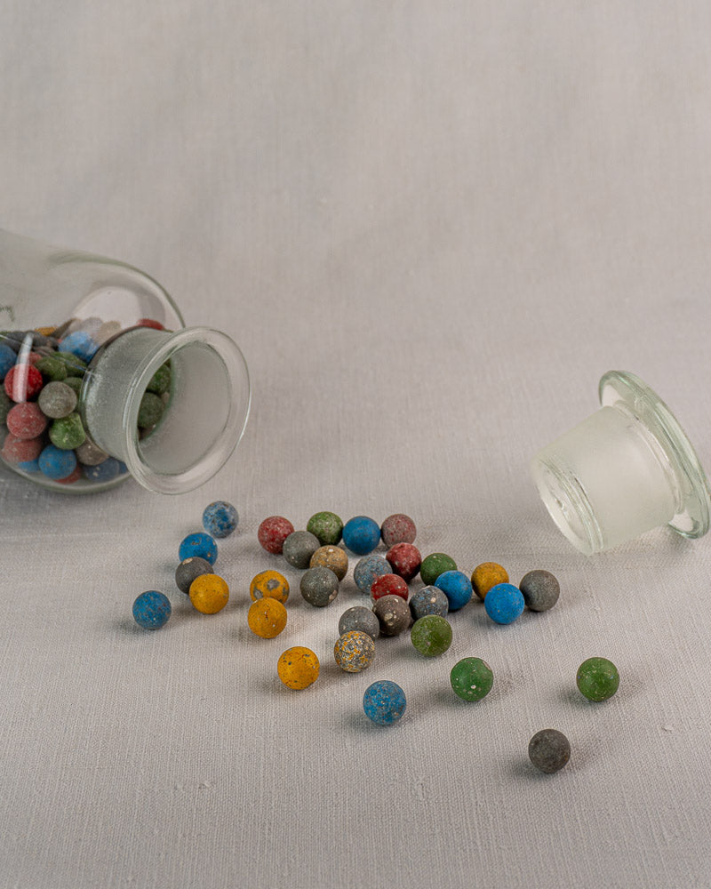 Antique French apothecary jar, stopper off, spills colorful clay marbles onto a light fabric surface.