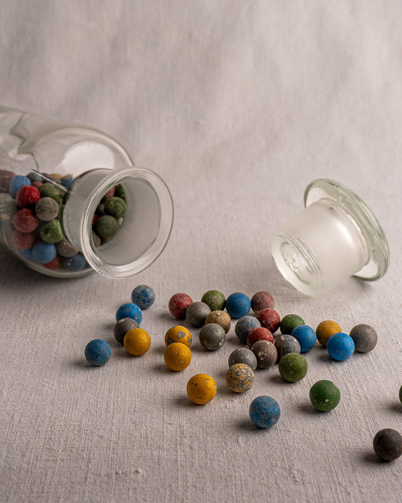 Antique French apothecary jar c. 1900 with marbles spilling onto light fabric, glass stopper beside it.