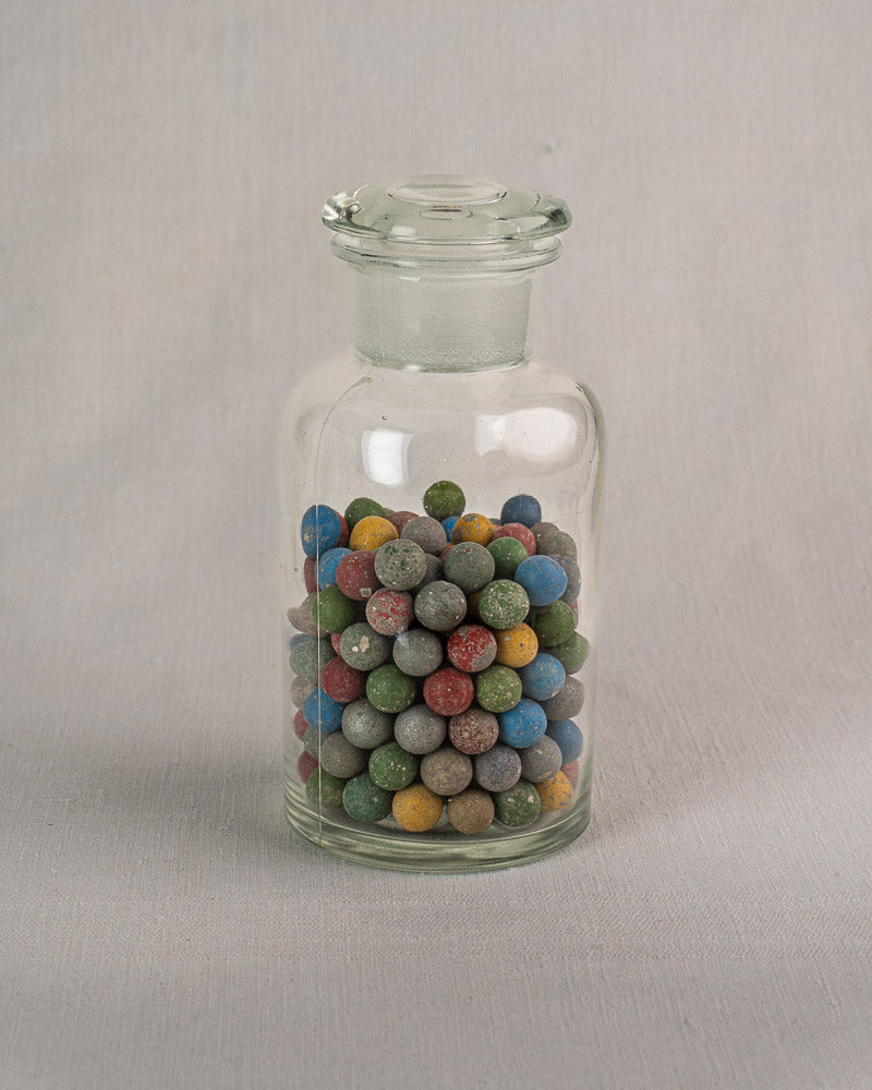 Antique French apothecary jar with stopper lid, filled with clay marbles, on light gray fabric surface.