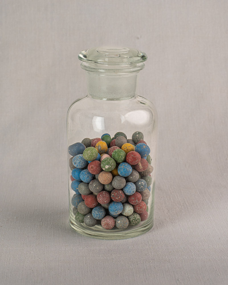 Antique French apothecary jar, clear hand-blown glass, filled with vintage clay marbles on a light gray surface.