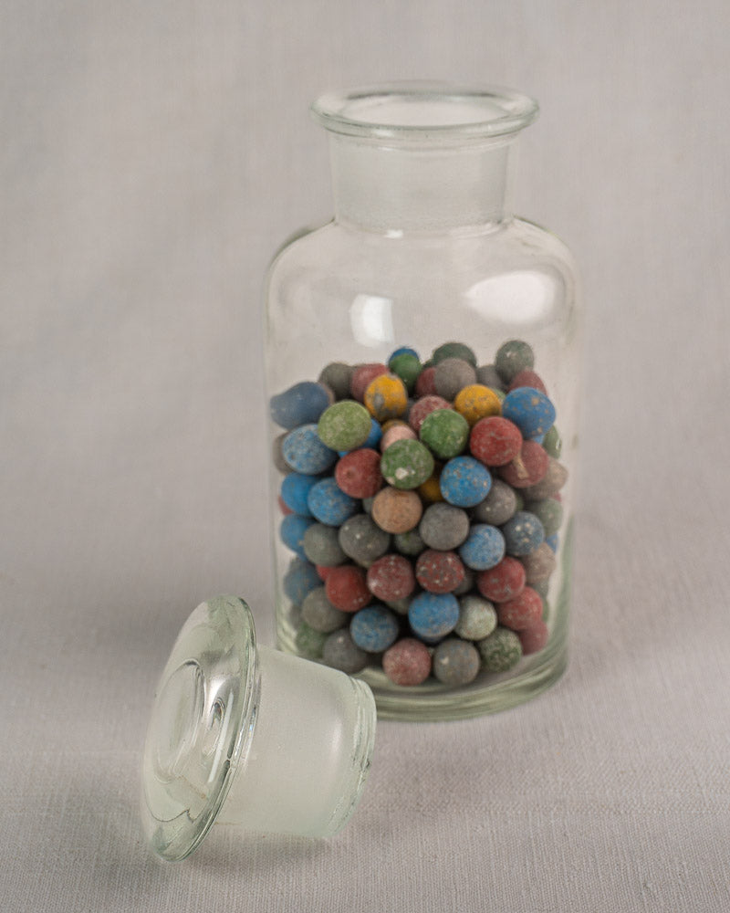 Antique French apothecary jar circa 1900 filled with clay marbles, glass lid resting beside on light surface.