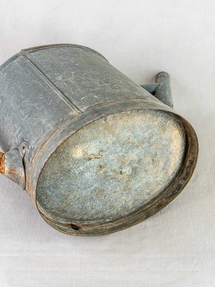 French zinc antique oval watering can shown on its side, neutral fabric backdrop.