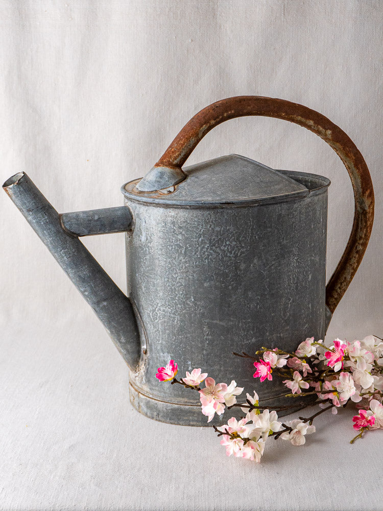 Antique French zinc oval watering can with rustic details, styled next to faux pink flowers.